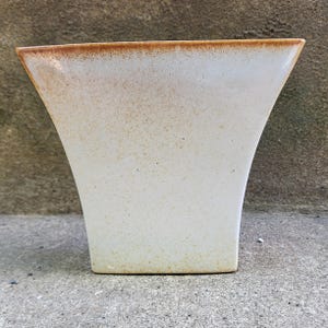 May include: A cream-colored ceramic vase with a flared, trapezoidal shape. The top edge has a brown, speckled rim. The vase has a speckled texture and a smooth, curved design. The vase is suitable for displaying flowers or as a decorative item.