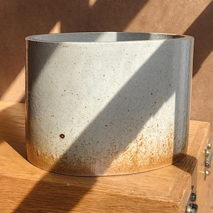 May include: A cylindrical, speckled ceramic planter with a light gray body and a brown, textured base. The planter sits on a wooden surface, with a shadow cast across its surface. The planter is suitable for indoor or outdoor use.
