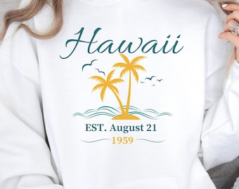 Puede incluir: Sudadera con capucha blanca con un gráfico de palmeras y el texto "Hawaii EST. August 21 1959" en amarillo y azul.