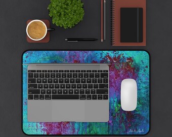 Op de afbeelding: Een computeropstelling met een laptop, muis en een kleurrijke muismat met abstracte kunst in blauw, paars en groen. Een koffiekopje, notitieblok en pen zijn ook aanwezig.