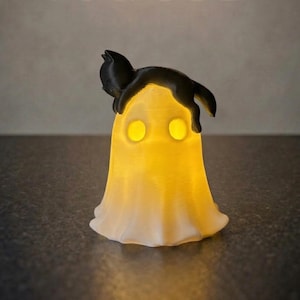 Könnte beinhalten: Eine Halloween-Themenlampe in Form eines Geistes mit einer schwarzen Katze obenauf. Der Geist hat zwei leuchtend gelbe Augen und wird von innen beleuchtet, wodurch ein sanftes, warmes Leuchten entsteht. Ein einzigartiger Dekorationsartikel.