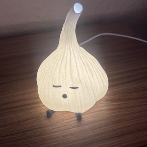 Könnte beinhalten: Eine skurrile Knoblauch-Nachtlampe, die sanft leuchtet. Die cremefarbene Lampe hat eine strukturierte Oberfläche, schwarze Füße und geschlossene Augen, was ihr einen schläfrigen Ausdruck verleiht. Der Stiel krümmt sich nach oben, mit hellem Licht an der Spitze.