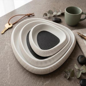 Könnte beinhalten: Drei weiße Keramikschalen mit geriffelter Außenseite, ineinander verschachtelt. Im Inneren befinden sich zwei schwarze, kieselsteinförmige Steine. Eine salbeigrüne Tasse, ein Schlüssel und ein Notizbuch sind ebenfalls zu sehen.