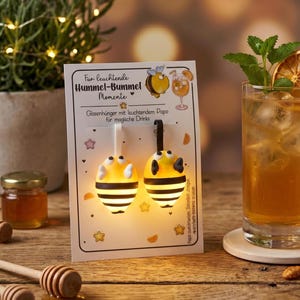 Könnte beinhalten: Zwei bienenförmige Glasanhänger auf einer weißen Präsentationskarte mit dem Text "Für leuchtende Hummel-Bummel Momente" und "Glasenhünger mit leuchtendem Popo für magische Drinks". Die Anhänger haben gelbe Körper mit schwarzen Streifen und weißen Flügeln.