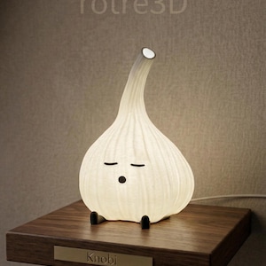 Könnte beinhalten: Eine skurrile, weiße Knoblauch-Lampe mit einem lächelnden Gesicht, schwarzen Füßen und einem gebogenen Hals. Die Lampe steht auf einem dunklen Holzregal mit dem Namen "Knobi" auf einem kleinen goldenen Schild. Die Lampe leuchtet und strahlt ein sanftes Licht aus.