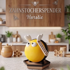 Op de afbeelding: Een grillige, geel en zwarte tandenstokerhouder in de vorm van een bij met witte antennes en een label met de tekst "Zahnstocher". De houder staat op een bruine zeshoekige basis. De achtergrond toont keukenspullen.