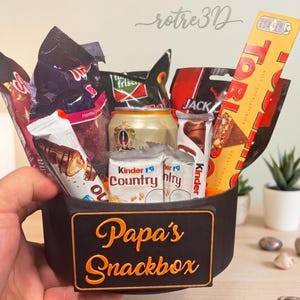 Könnte beinhalten: Eine schwarze, 3D-gedruckte, runde Snackbox mit einem orangefarbenen Schild, auf dem "Papa's Snackbox" in orangefarbenen Buchstaben steht. Der Behälter ist mit verschiedenen Snacks gefüllt, darunter ein Toblerone-Riegel, Kinder Country-Riegel, eine Getränkedose und verschiedene Süßigkeitentüten.