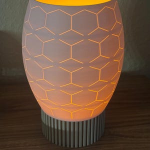 Op de afbeelding: Een witte keramische tafellamp met een geometrisch honingraatpatroon. De lamp straalt een warme, gele gloed uit. De basis is lichtgrijs met verticale lijnen. De lamp staat op een houten oppervlak.