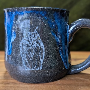 May include: A handmade ceramic mug with a dark speckled finish and a blue glaze dripping down the sides. A white outline of an owl is featured on the front. The mug has a large, rounded handle.