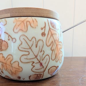 May include: A round ceramic jar with a light cream-colored glaze featuring a brown hand-drawn pattern of oak leaves and acorns. The jar has a flat, circular bamboo lid and a thin, silver-colored metal spoon resting inside the top opening.
