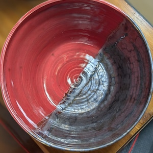 May include: A handmade ceramic bowl with a striking two-tone design. The left half is a vibrant red, while the right half transitions to a dark, textured gray. The bowl's interior features concentric circles, showcasing the pottery's craftsmanship.