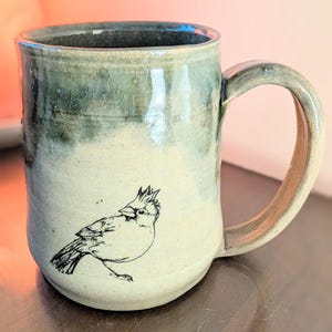 May include: A ceramic mug with a two-tone glaze featuring a dark green top section and a light beige bottom section. A black line drawing of a crested bird is printed on the lower front of the mug, which includes a curved handle on the right side.