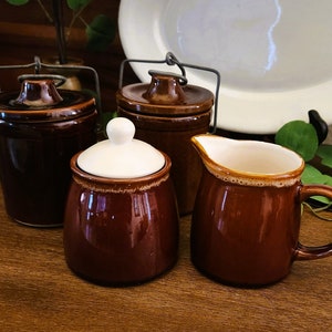 Peut inclure: Un ensemble de quatre articles de cuisine en céramique brune, comprenant deux pots à couvercle avec des poignées en fil de fer, un sucrier avec un couvercle blanc et un pot à crème.