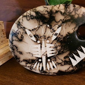 Puede incluir: Un jarrón de cerámica blanco y negro con un diseño de mariposa. El jarrón tiene una superficie texturizada y un agujero en el lateral para flores.