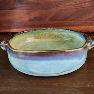 Puede incluir: Un pequeño plato para hornear de cerámica de forma ovalada con dos asas. El plato está esmaltado en tonos de verde, marrón y azul.