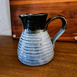 Puede incluir: Jarra de cerámica hecha a mano con borde y asa negros, que transiciona a un cuerpo a rayas azules y blancas. La jarra tiene un pico para verter fácilmente y un asa resistente. Esta pieza única es perfecta para servir bebidas.