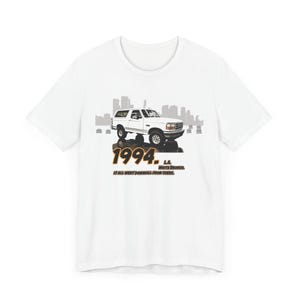 Puede incluir: Camiseta blanca con un gráfico de un Ford Bronco blanco de 1994 frente a un horizonte urbano. El texto "1994. L.A. White Bronco. It all went downhill from there." está impreso debajo de la imagen.