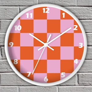 May include: A round wall clock with a white frame. The clock face features a checkerboard pattern of orange and pink squares. White numerals mark the hours, and white hands indicate the time.