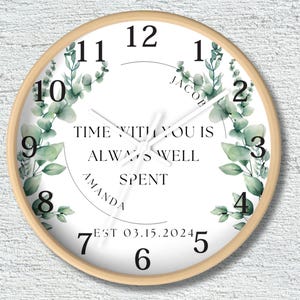 May include: Round wall clock with a light wood frame. The clock face is white with black numerals and hands. It features the text "TIME WITH YOU IS ALWAYS WELL SPENT" and decorative eucalyptus. The names "JACOB" and "AMANDA" are also included.