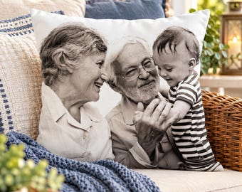 May include: A square white throw pillow featuring a black and white printed photograph of an older woman, an older man with glasses and a beard, and a young child wearing a striped shirt, all smiling and interacting together on the cushion.