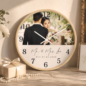 May include: A round wall clock with a light wood frame featuring a custom photograph of a couple on the upper half and the text Mr. & Mrs. Lee EST 01.24.2026 printed on the lower white half with black numbers from 1 to 12 and white clock hands.