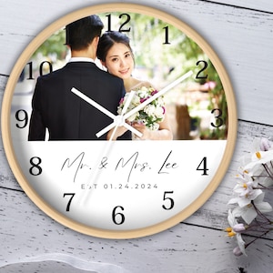 May include: A white round clock with a wood frame. The clock face features a wedding photo of a couple with a bouquet of flowers. The text "Mr. & Mrs. Lee EST 01.24.2024" is printed below the photo.