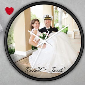 May include: Circular black wall clock displaying a wedding photograph of a couple. The image shows a bride in a white gown and a groom in a navy uniform. The clock hands are white, and the names "Rachel + Jacob" are written on the clock face.