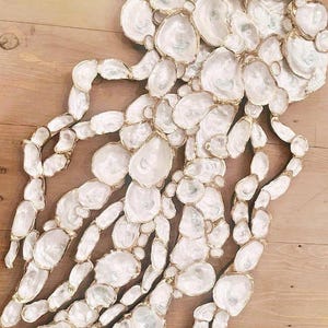 May include: A decorative wall art piece made of numerous white oyster shells with gold-colored edges. The shells are arranged in a jellyfish-like shape, creating a unique and elegant design. The artwork is placed on a wooden surface.