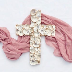 May include: A decorative cross made of oyster shells, with a gold trim, resting on a pink fabric. The cross is centered in the image, with the pink fabric draped around it on a white background. The oyster shells have a pearlescent sheen.