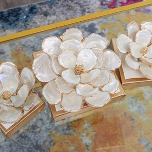 May include: Three white flower sculptures made of oyster shells with gold accents on a gold base. The flowers are arranged on a glass surface with a colorful rug underneath.