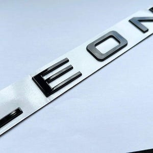 May include: Black "LEON" emblem on a white backing. The letters are in a bold, sans-serif font. The emblem is likely for a car or vehicle.