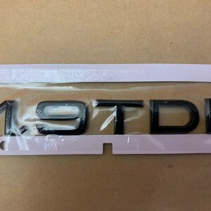 May include: Black 1.9 TDI badge on a white backing. The badge is rectangular with rounded corners. The letters and numbers are in a bold, sans-serif font.