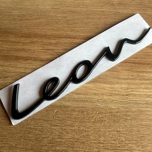 May include: A black "Leon" sticker in a cursive font, presented on a white backing. The sticker is set against a wooden surface. The glossy finish of the sticker contrasts with the matte paper.