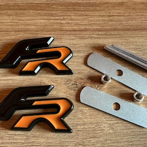 May include: Two black and orange FR emblems with a glossy finish, accompanied by two silver metal mounting plates, two small hexagonal nuts, and two threaded metal rods for installation, all displayed as a set of automotive exterior accessories.