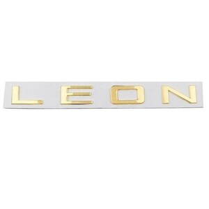 May include: Gold-coloured metal lettering spelling out the word "LEON" on a white rectangular backing. The letters are in a sans-serif font and appear three-dimensional, with subtle shadows.