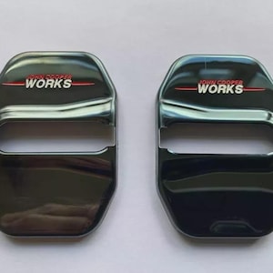 May include: Two black car door lock covers with the text "JOHN COOPER WORKS" in white and red. The covers have a glossy finish and a rectangular shape with a curved top and a slot in the middle.