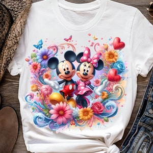 May include: White t-shirt with a colorful graphic print of Mickey Mouse and Minnie Mouse surrounded by flowers and hearts.