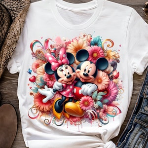 May include: White t-shirt with a colorful graphic print of Mickey Mouse and Minnie Mouse surrounded by flowers.