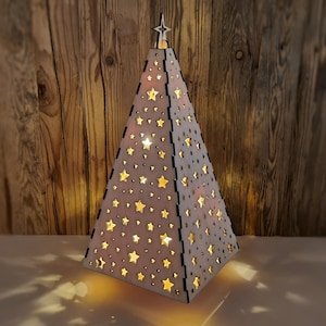 Puede incluir: Un árbol de Navidad de madera en forma de pirámide con recortes de estrellas. El árbol está iluminado desde dentro, proyectando sombras en forma de estrella. Una estrella corona el árbol. El árbol mide unos 30 cm de alto.