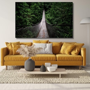 May include: A mustard yellow sofa with several matching cushions and one white cushion, featuring a wall art print of a suspension bridge in a forest. A gray vase with dried pampas grass and small white containers sit on a wooden coffee table.