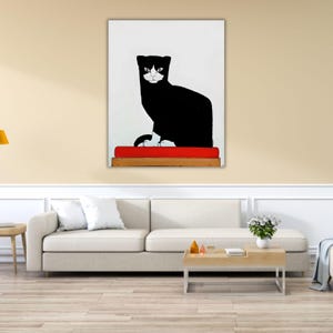 May include: A framed wall art print featuring a stylized black and white cat sitting on a bright red rectangular cushion. The cat has a solid black body with white markings on its face and paws, set against a plain white background.