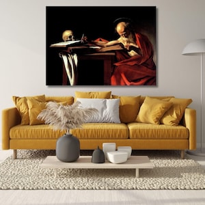 May include: A rectangular wall art print depicting a bearded figure in a red robe writing at a wooden desk with a skull nearby, displayed above a mustard yellow sofa with several throw pillows, a coffee table, and a floor lamp.