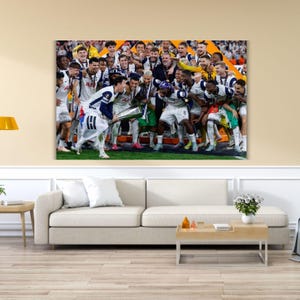 May include: A large rectangular wall art print depicting a group of soccer players in white and navy blue uniforms celebrating a victory on a field. The players are gathered together, cheering and holding a silver trophy cup aloft in the center.