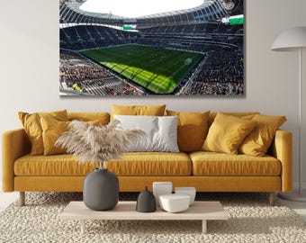 May include: A rectangular wall art print showing a high-angle view of a large football stadium with a bright green pitch, dark seating areas, and a modern roof structure, mounted on a wall above a mustard yellow sofa with various cushions.