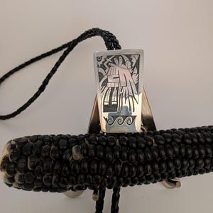 May include: A silver bolo tie with a rectangular pendant featuring an etched design of a figure and geometric shapes. The bolo tie has a black braided cord and is displayed on a black corn cob.