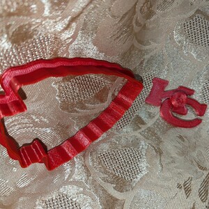 May include: A red heart-shaped cookie cutter and the letters "KC" in a cursive font. The cookie cutter has a ridged edge and is made of a solid red material. The letters are also red and appear to be made of the same material.