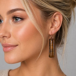 May include: A woman wearing a pair of rectangular, brown and gold earrings. The earrings are hanging from copper-colored hooks. The earrings have a striped pattern and are a long, vertical shape.