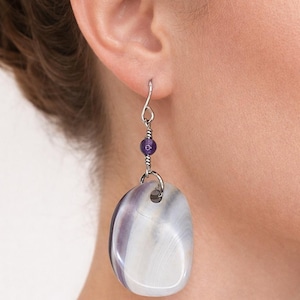 May include: A silver earring with a large, oval, polished stone in shades of white and purple. A small, round, purple bead is suspended above the stone, connected by silver wire. The earring is designed to be worn in the ear.