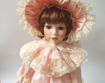 May include: A porcelain doll with auburn hair, blue eyes, and painted lips. The doll wears a light pink satin dress with a ruffled collar and a matching wide-brimmed bonnet, both trimmed with intricate cream-colored lace and a white ribbon tie.