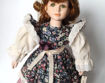 May include: A porcelain doll with auburn hair and green eyes, wearing a dark blue dress with a multi-colored floral pattern, white puffed sleeves with eyelet lace trim, a cream-colored ribbon at the waist, and a tag labeled EMILY.